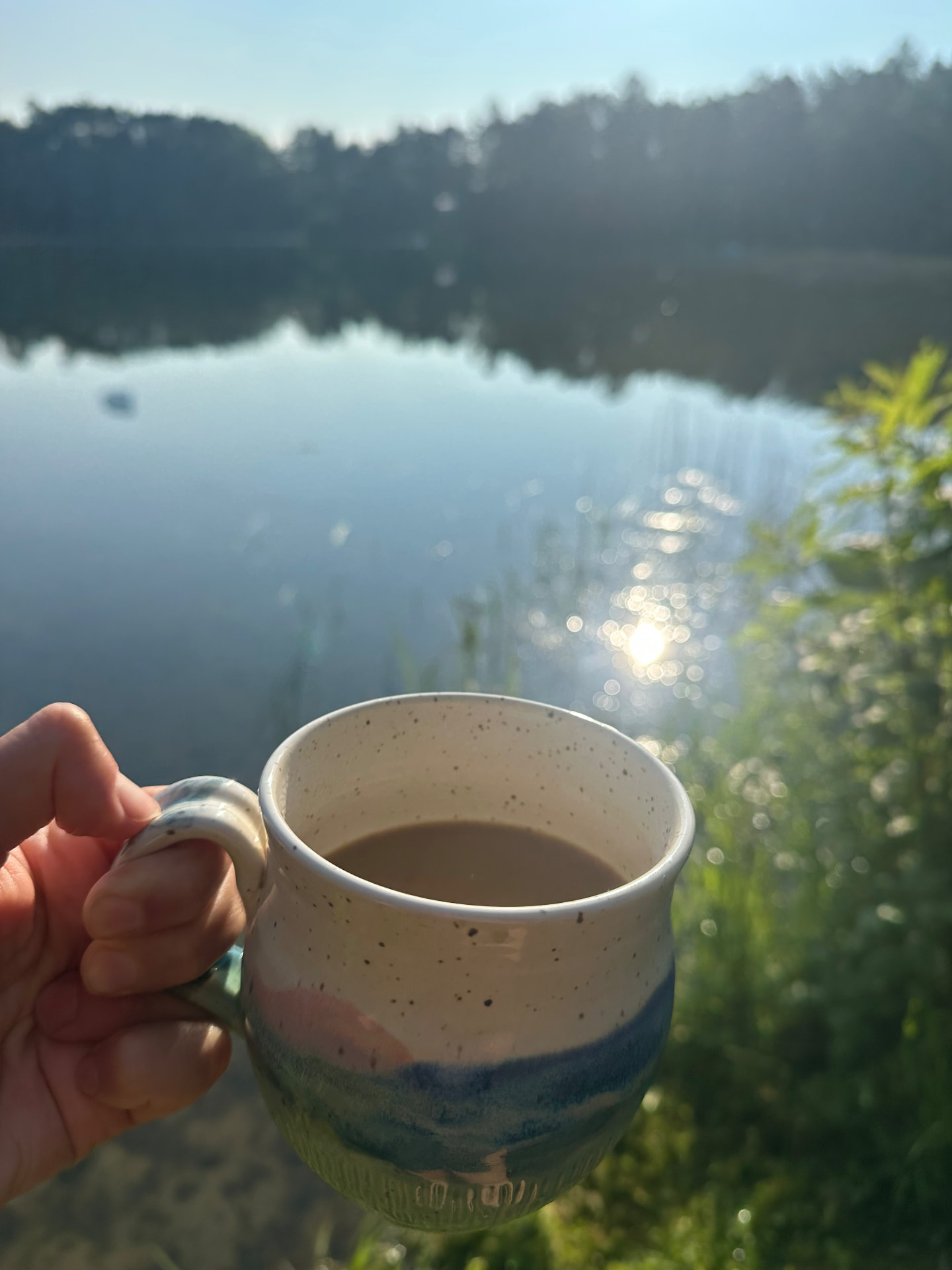 Hand holding a pottery mug of coffee overlooking a calm lake at the cottage in the morning sun