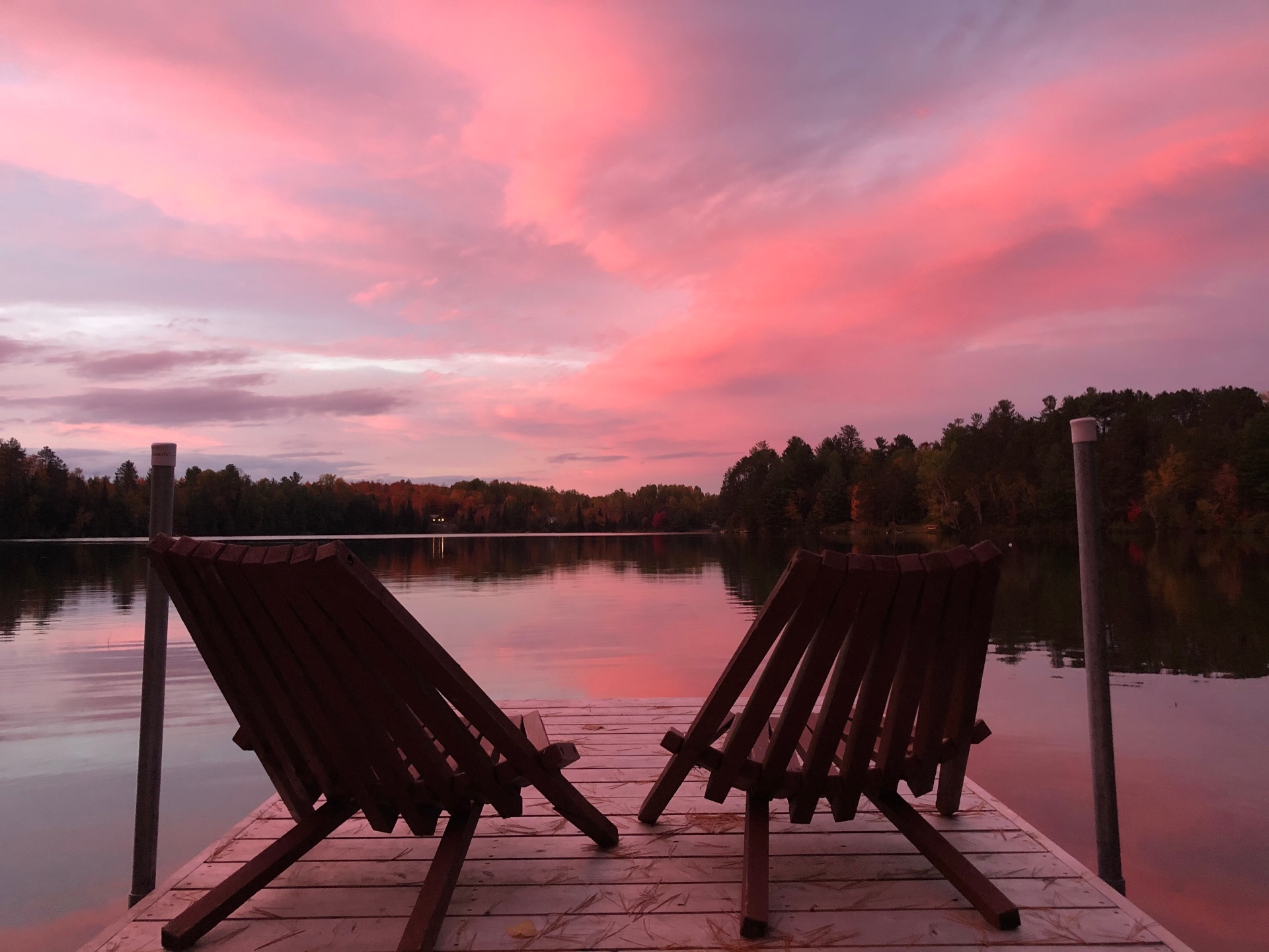 Two Muskoka chairs on a dock at pink sunset over a calm Canadian lake